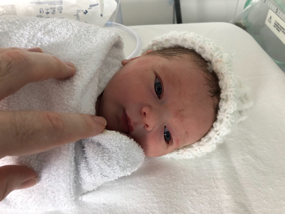 Baby Max, a few minutes after birth, in an incubator wrapped in a towel and wearing a fluffy hat. He's looking calmly toward the camera.